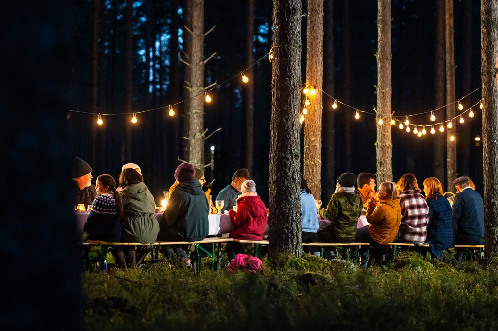 Ihmisiä istuu yhdessä ison ruokapöydän ääressä metsässä.