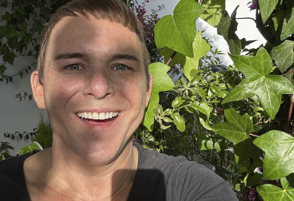Photographer Kristian Kaarna in his own garden. Ivy leaves filter sunlight onto his face, creating a warm and relaxed atmosphere. The image reflects the humanity and positive spirit of the SmilingOulu26 project.