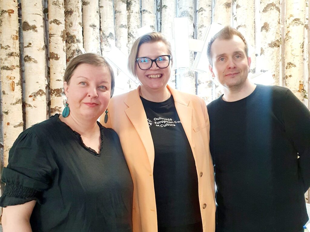 Kolme henkilöä hymyilee tilassa, joka on koristeltu koivuille